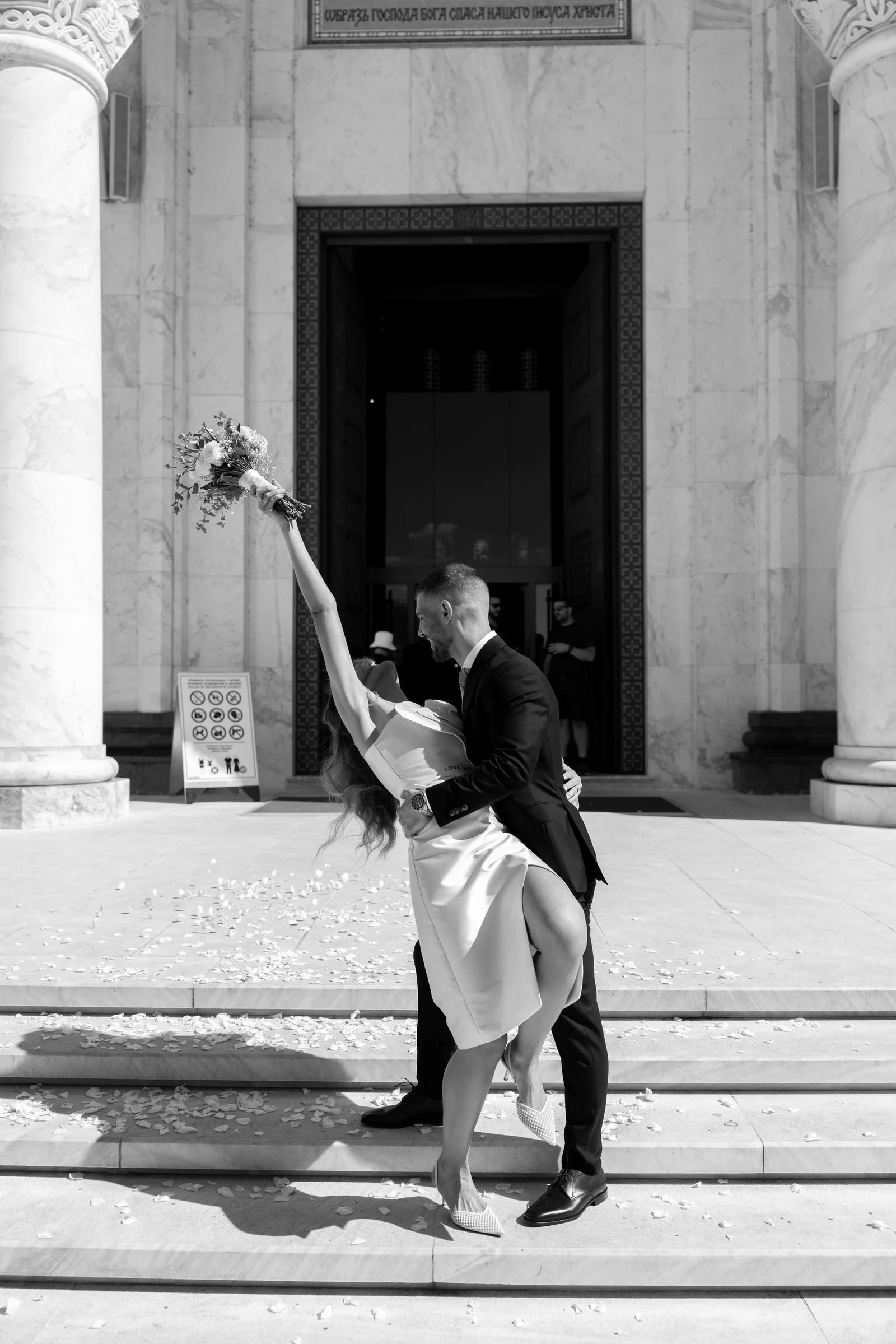 Groom dips the bride in a joyful dance on the marble church steps scattered with petals.