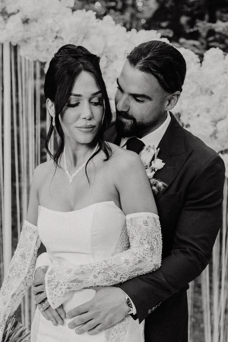 Groom embraces a bride in lace sleeves from behind in front of a white orchid backdrop