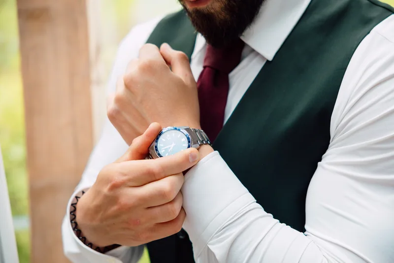 Close-up of a groom in a green vest and burgundy tie fastening a silver wristwatch