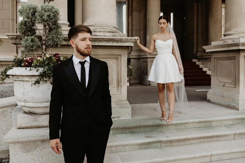 A groom stands in the foreground while the bride waits on stone steps in a short dress and long veil.