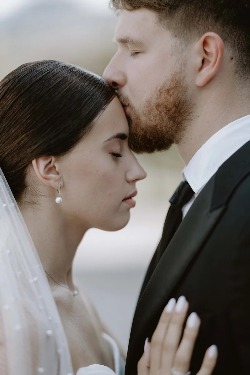 A groom tenderly kisses the bride's forehead as she closes her eyes, a pearl earring catching the light.