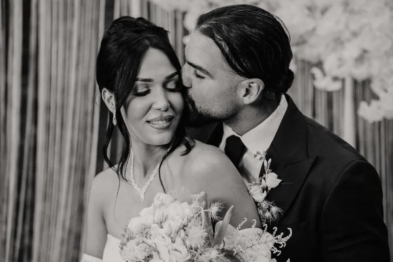 Black and white image of a groom kissing the temple of a smiling bride holding her bouquet