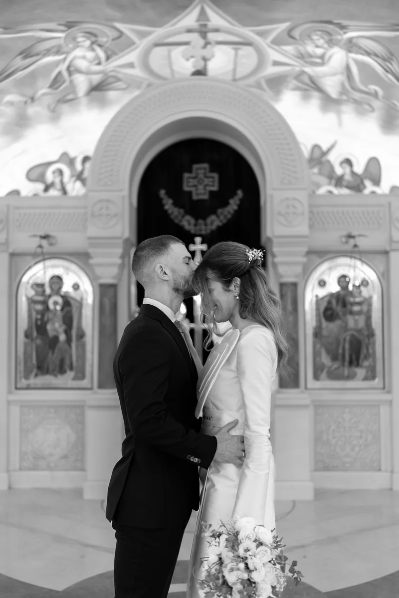 Groom kisses the bride's forehead before a carved iconostasis topped with painted angels.