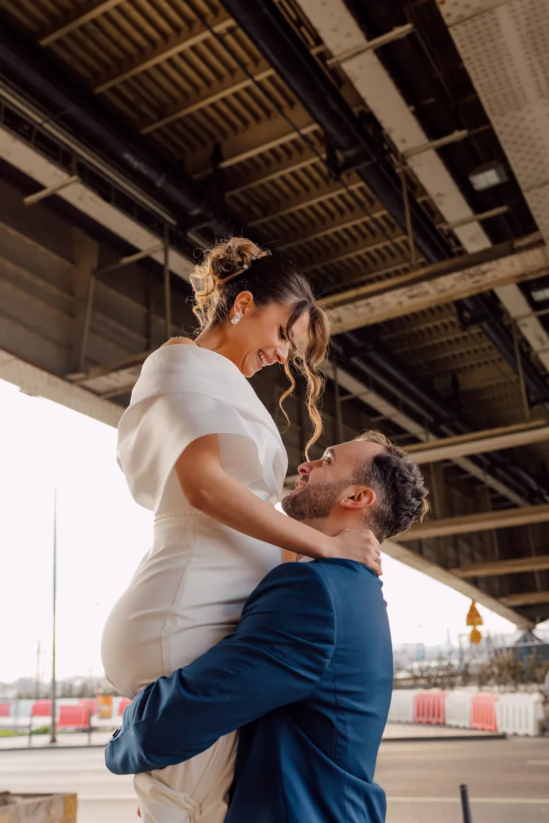 A groom lifts his laughing bride into the air beneath the steel underside of a city bridge.
