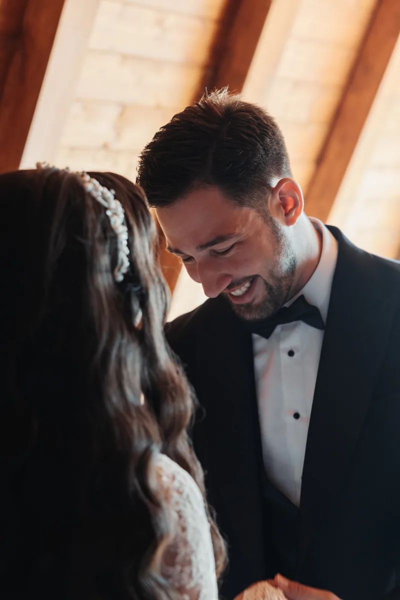 Groom in a bow tie tuxedo smiles warmly down at his bride in soft golden light.