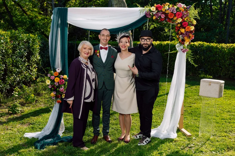 Four people pose together under a draped floral garden arch with bright orange and purple blooms