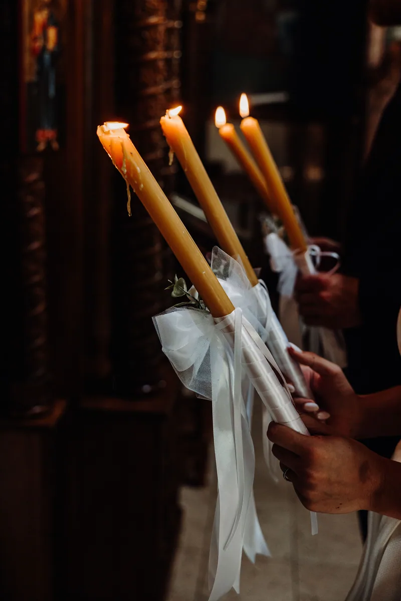 Hands hold tall lit ceremony candles wrapped in tulle and ribbon inside a warmly lit church.
