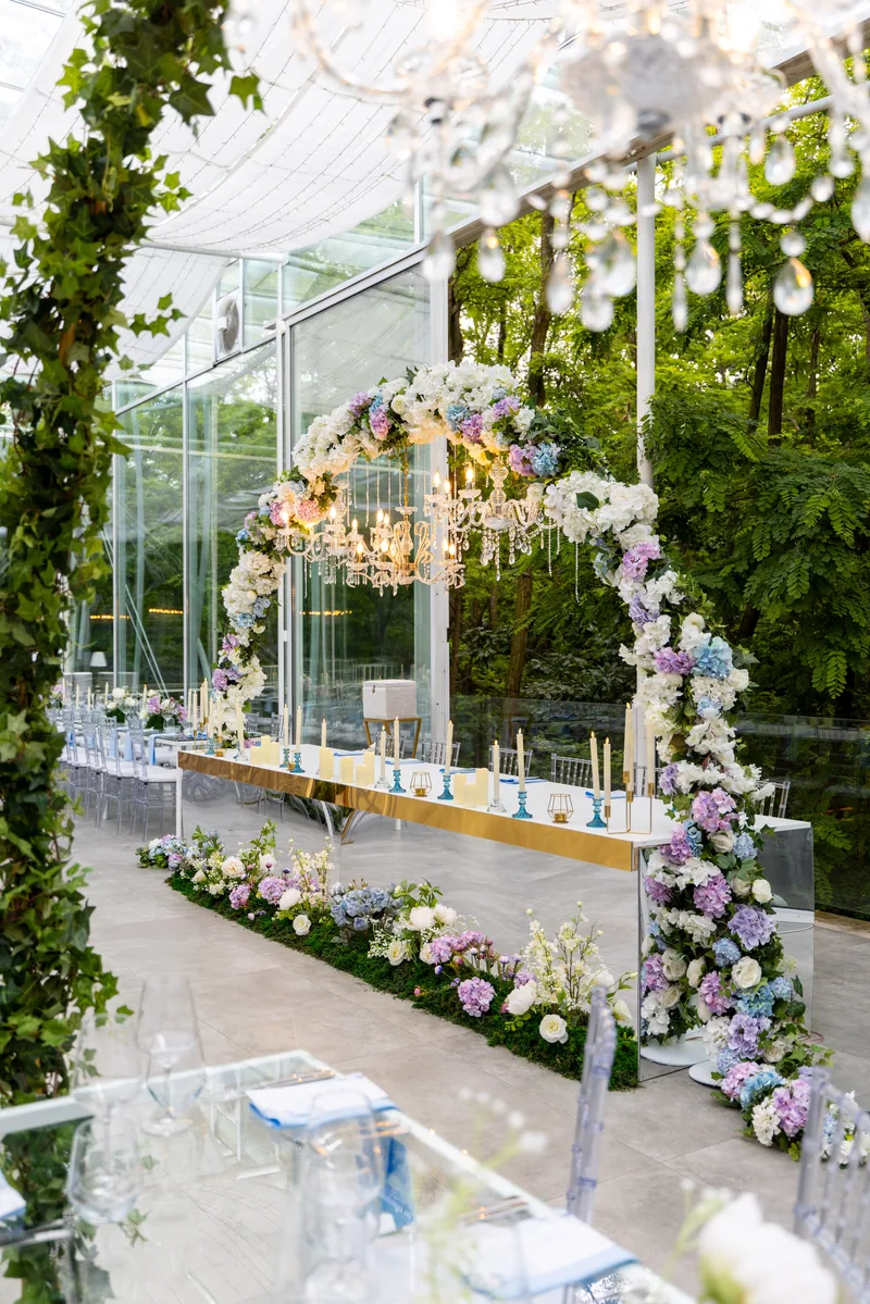 Long head table with a hydrangea arch, tall candles and a crystal chandelier in a glass pavilion