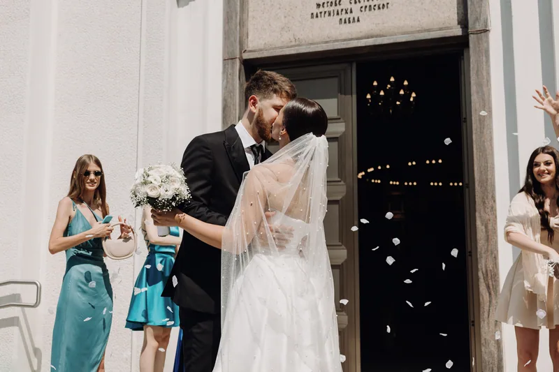 A couple kiss in a sunny doorway as guests shower them with flower petals, the bride holding white roses.