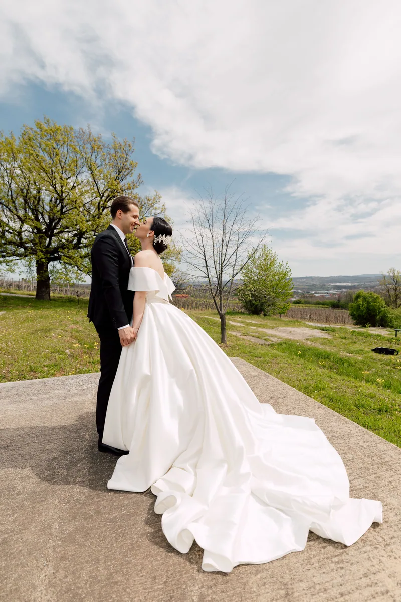 A couple kiss on an overlook as the bride's long satin train spreads across the ground above the vineyards.