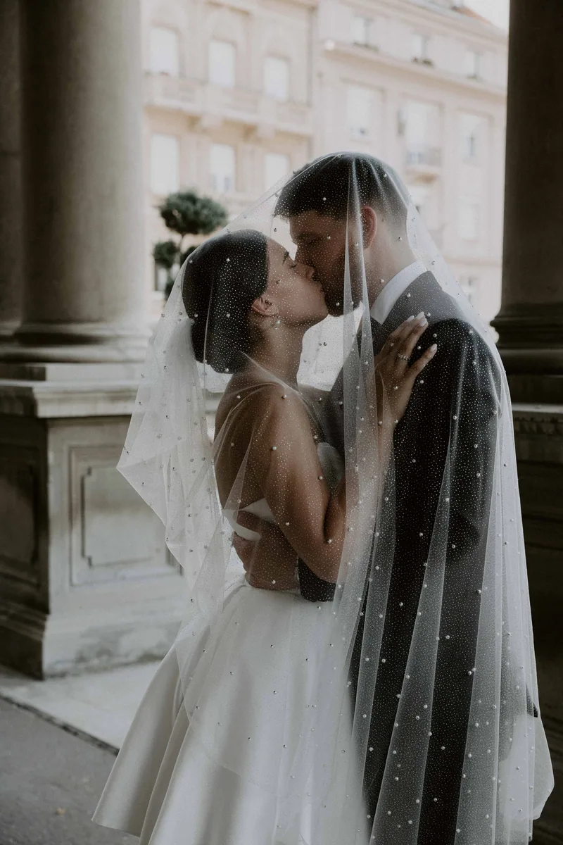 Newlyweds share a kiss under a pearl dotted veil among pale stone columns with a topiary tree behind.