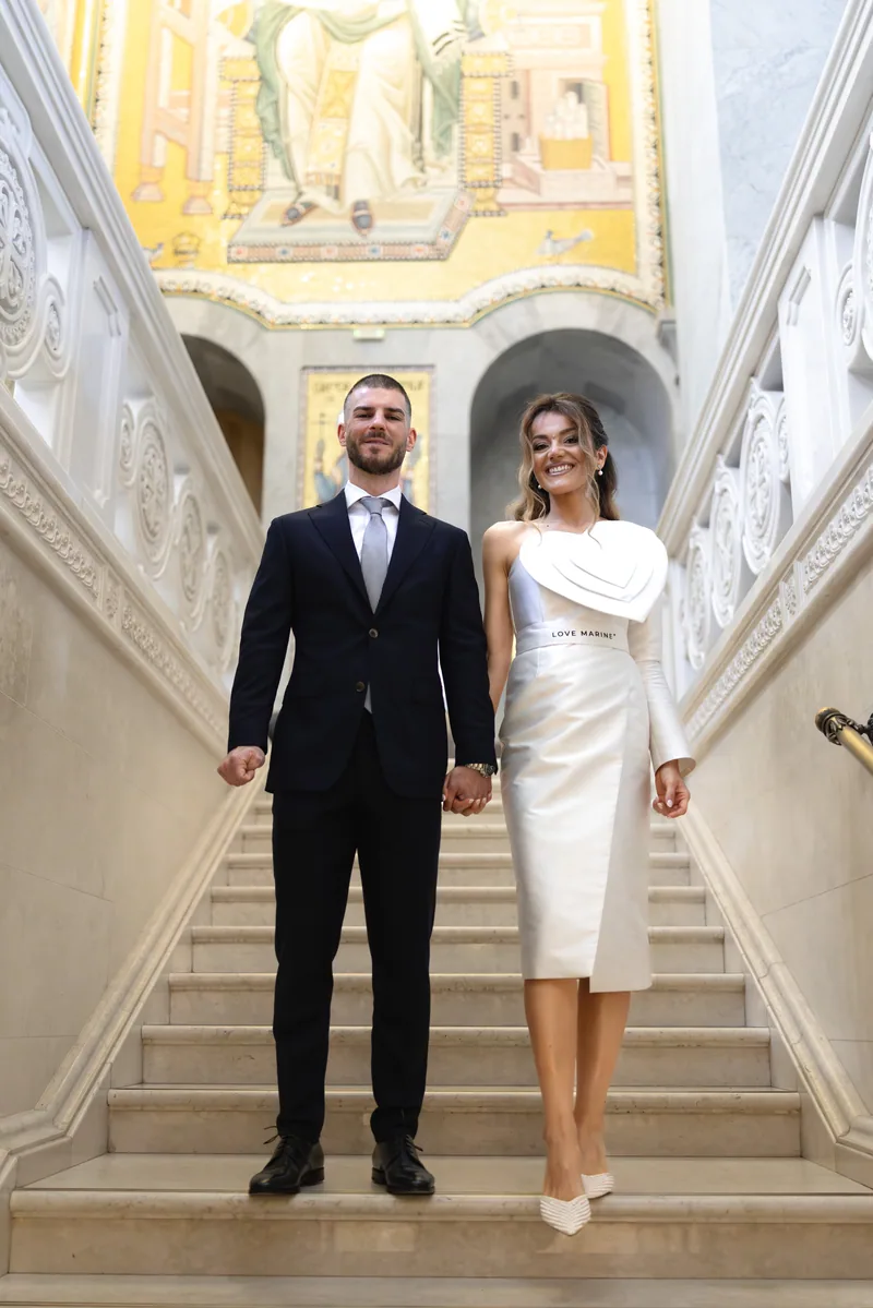 Newlyweds hold hands on a marble stairway with a glowing golden mosaic saint behind them.