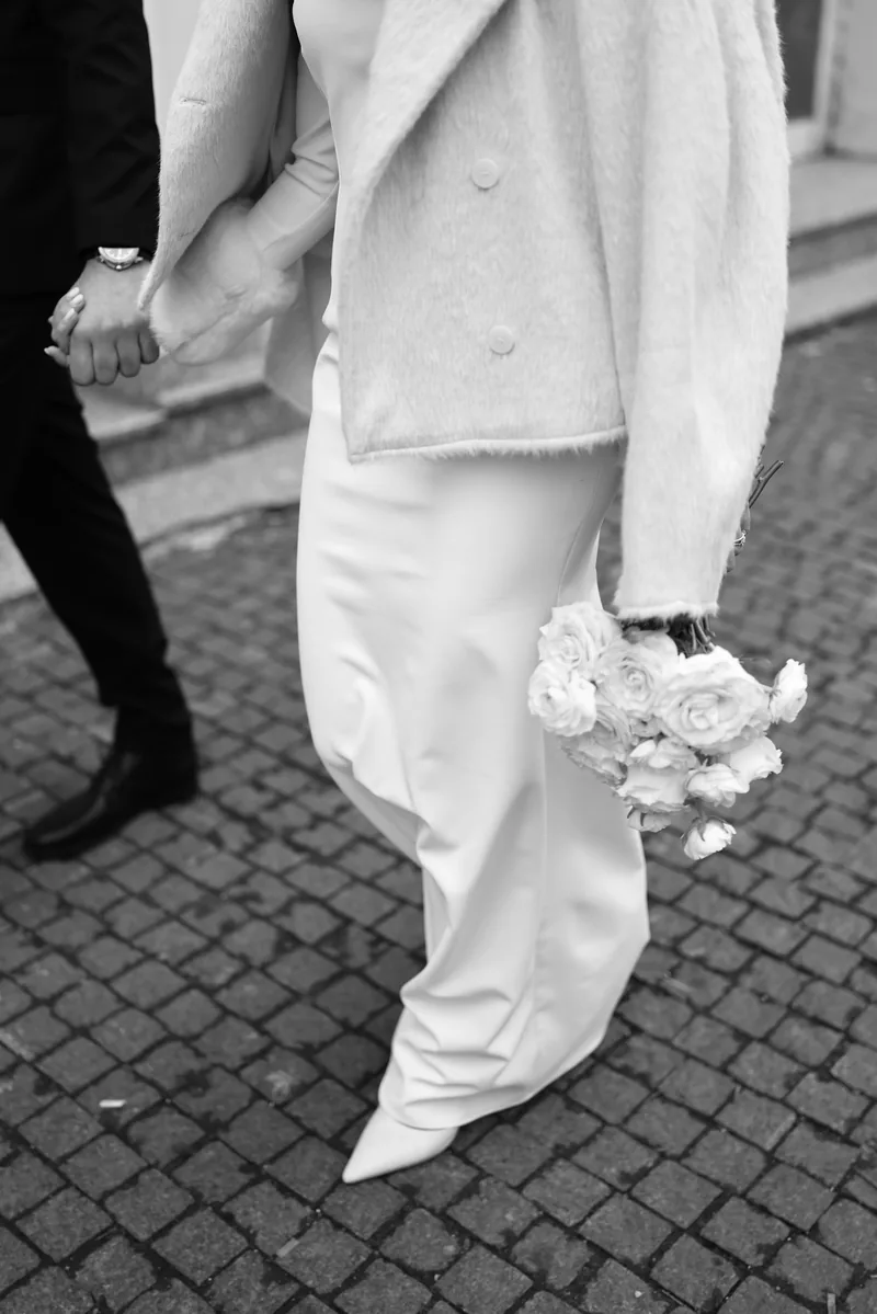 In black and white, a couple walk hand in hand over cobblestones as the bride carries a white rose bouquet.