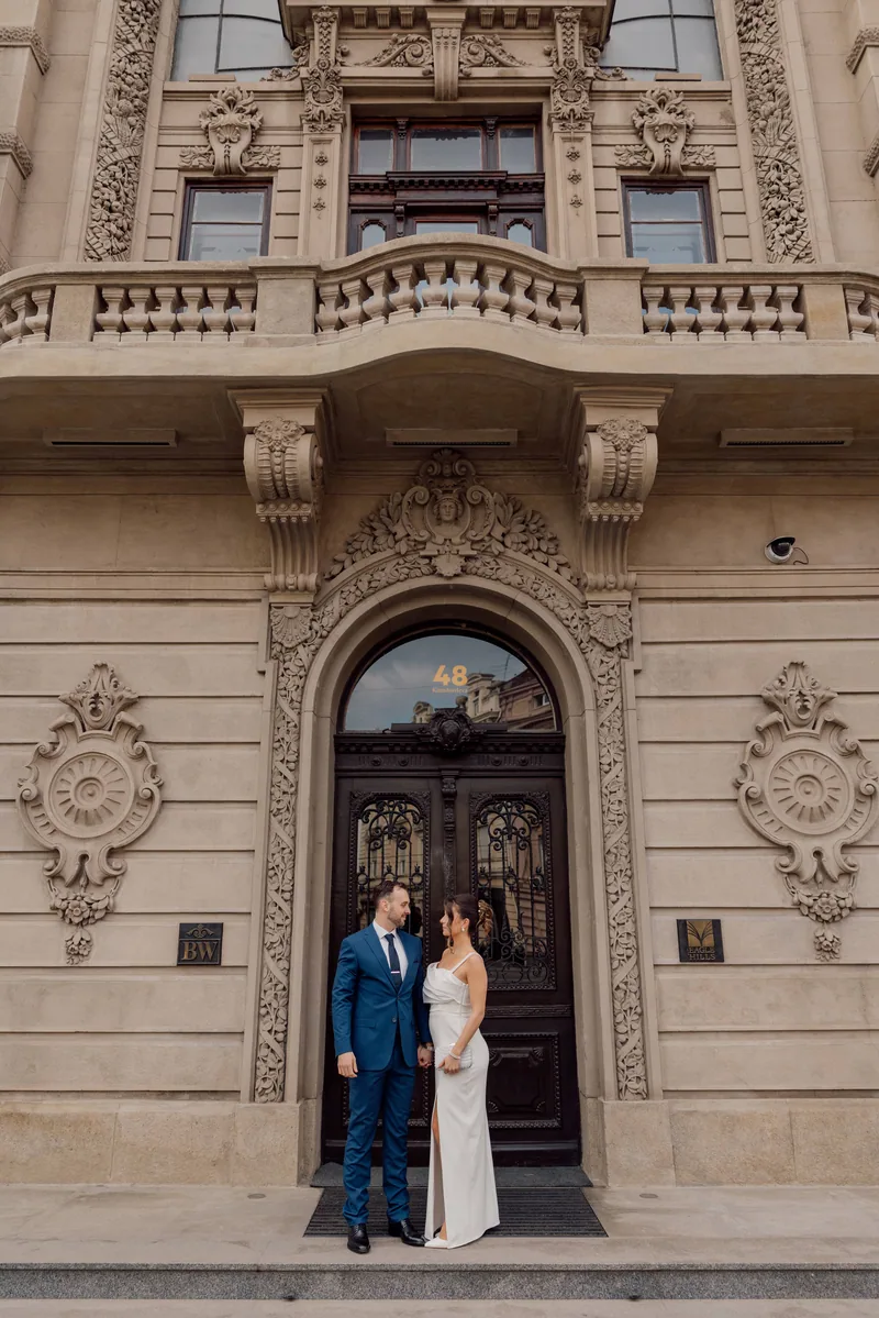 A bride and groom hold hands before an ornate carved Baroque doorway of a historic city building.