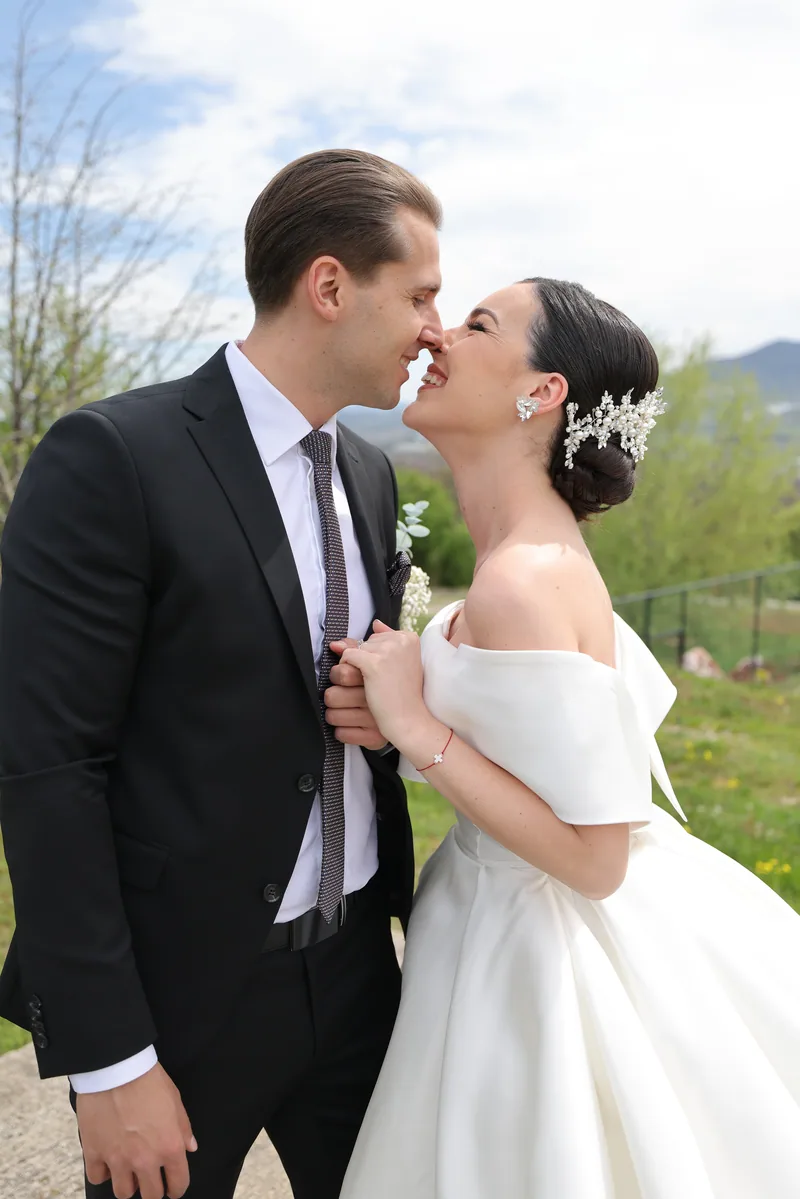 A bride in an off-shoulder ballgown and her groom laugh nose to nose on a green spring hilltop.
