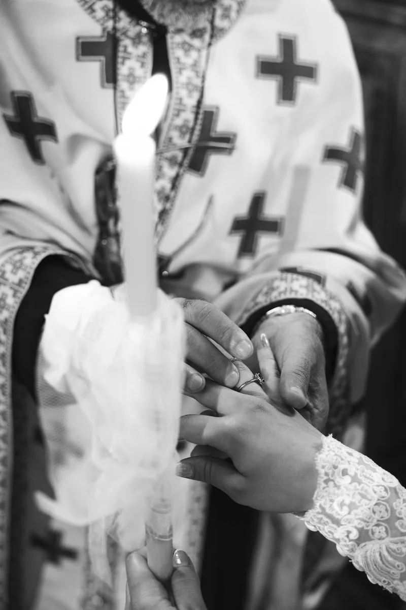 Priest blesses the ring on the bride's hand beside a tall lit ceremony candle.
