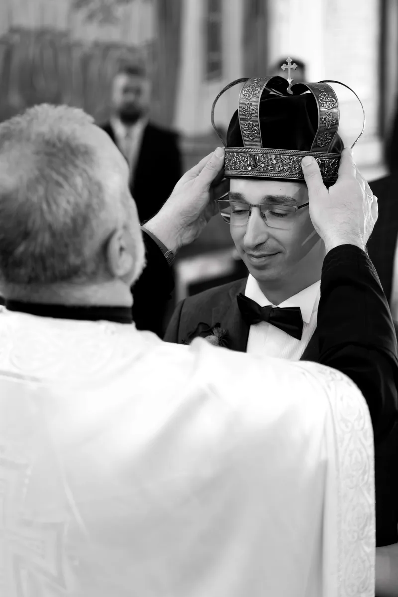 Black and white moment of a priest holding a ceremonial crown above a smiling groom's head