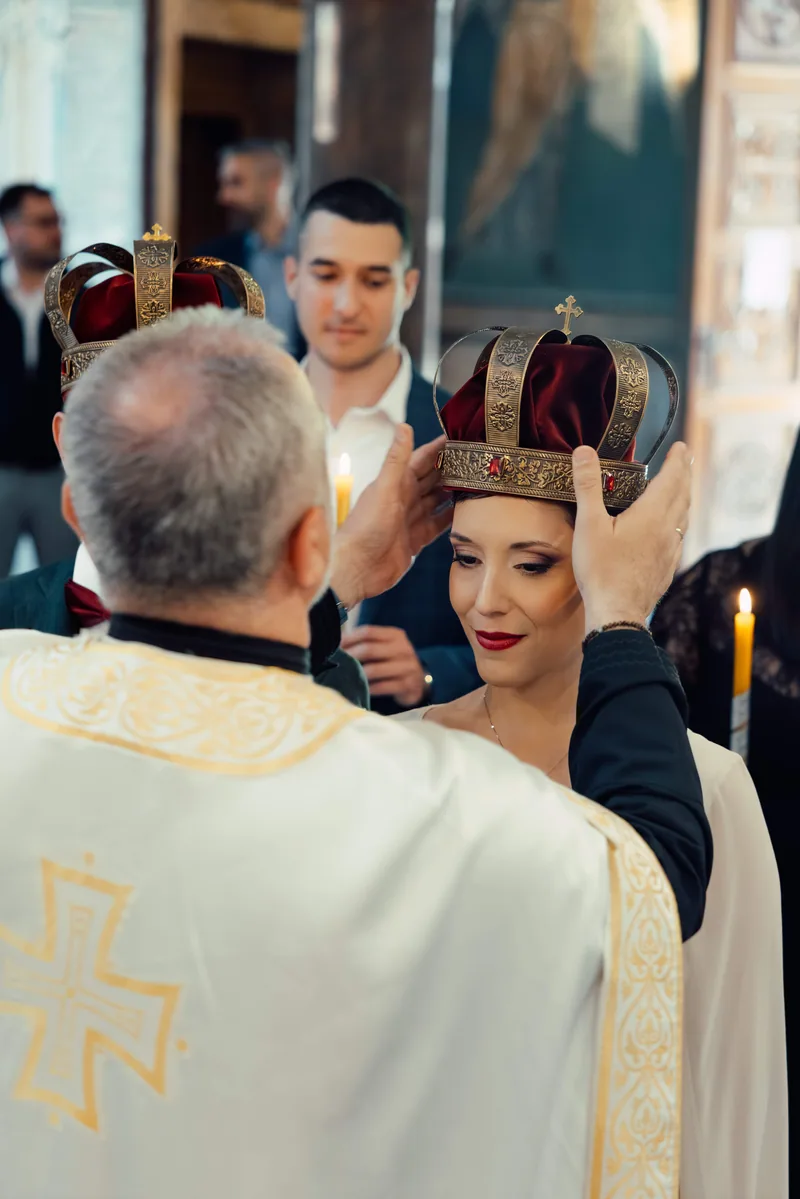 A priest lowers an ornate gold and velvet crown onto the head of a smiling bride in the church