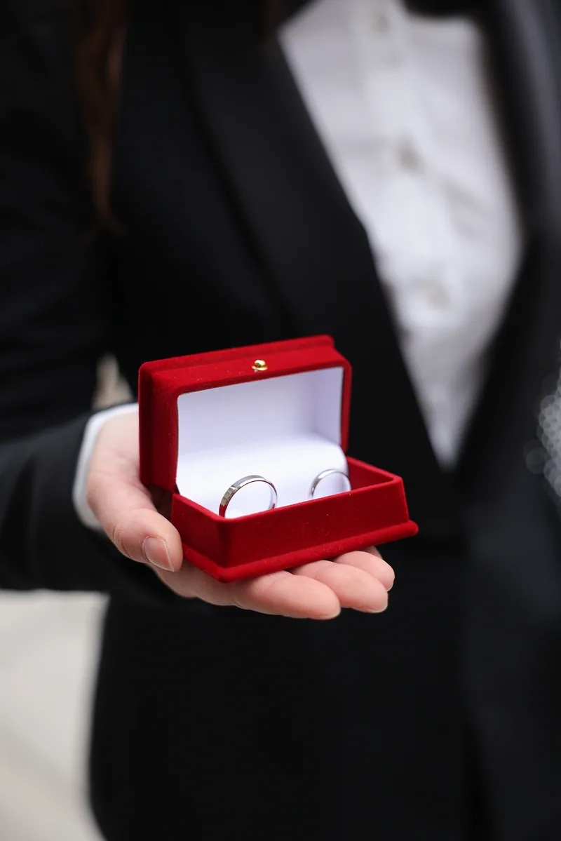 A hand holds an open red velvet box displaying two silver wedding bands against a black tuxedo.