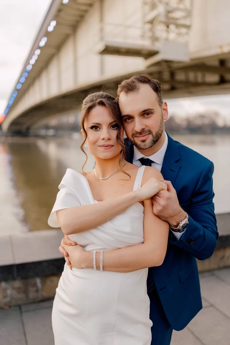 A bride and groom look to the camera in a close embrace with a lit bridge and river behind them.