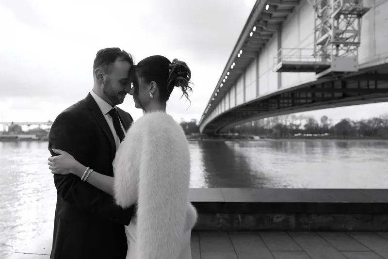 In black and white, a couple touch foreheads by a river railing as a lit bridge stretches across the water.