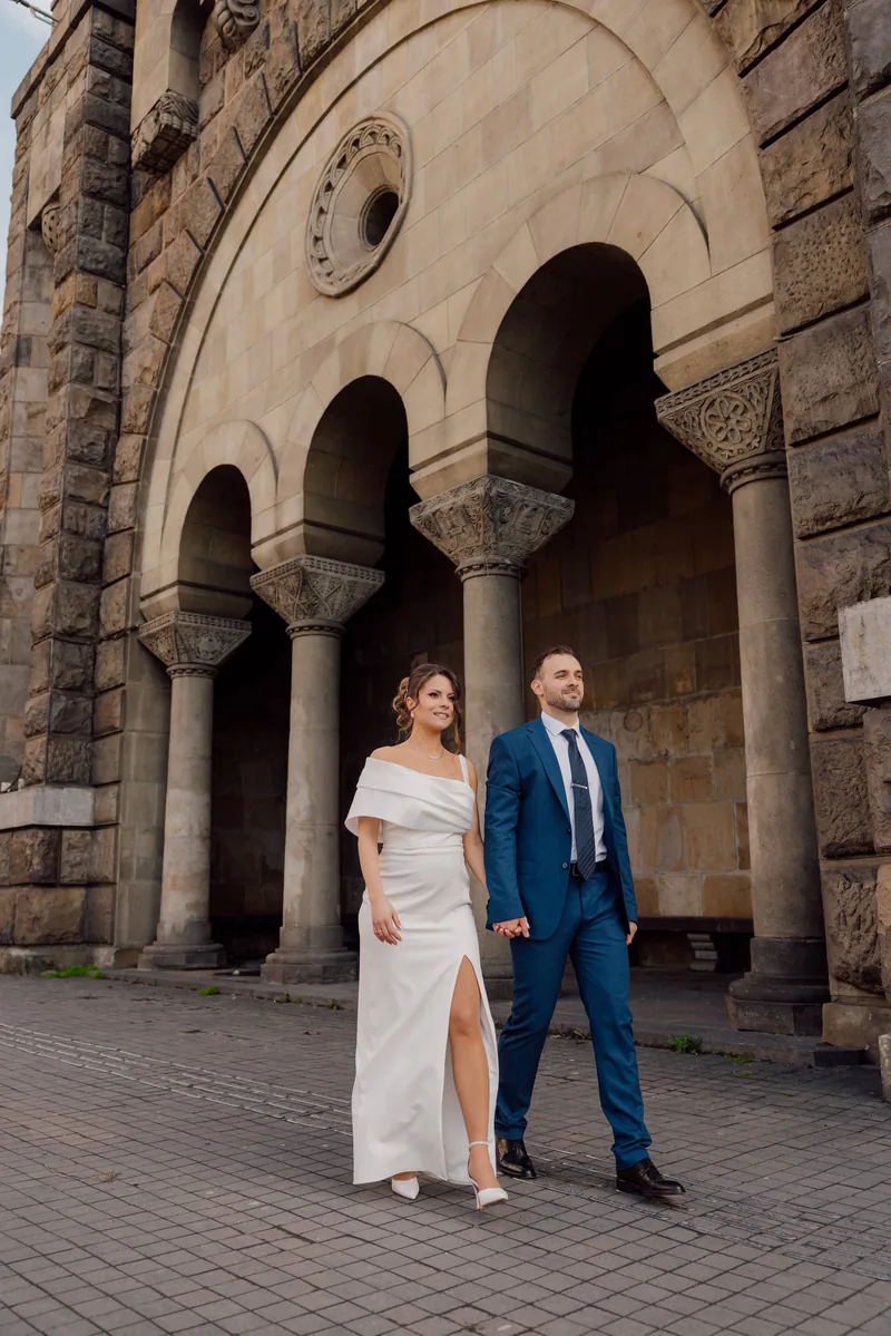 A bride in a slim white dress and a groom in a blue suit walk hand in hand before a grand arched stone facade.
