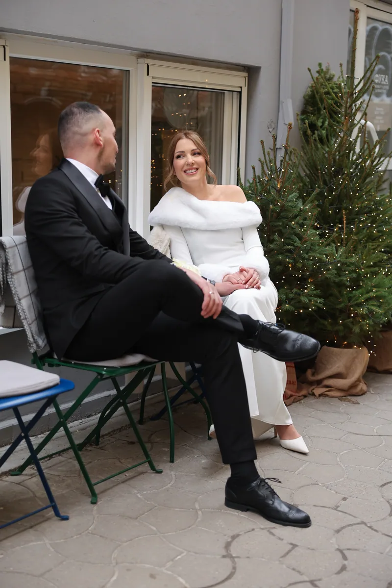 A laughing bride and her groom sit on bistro chairs on a terrace beside a lit Christmas tree.