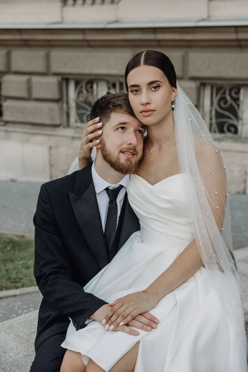 A bride sits on her groom's lap on stone steps, her hand at his cheek, wearing a long pearl dotted veil.