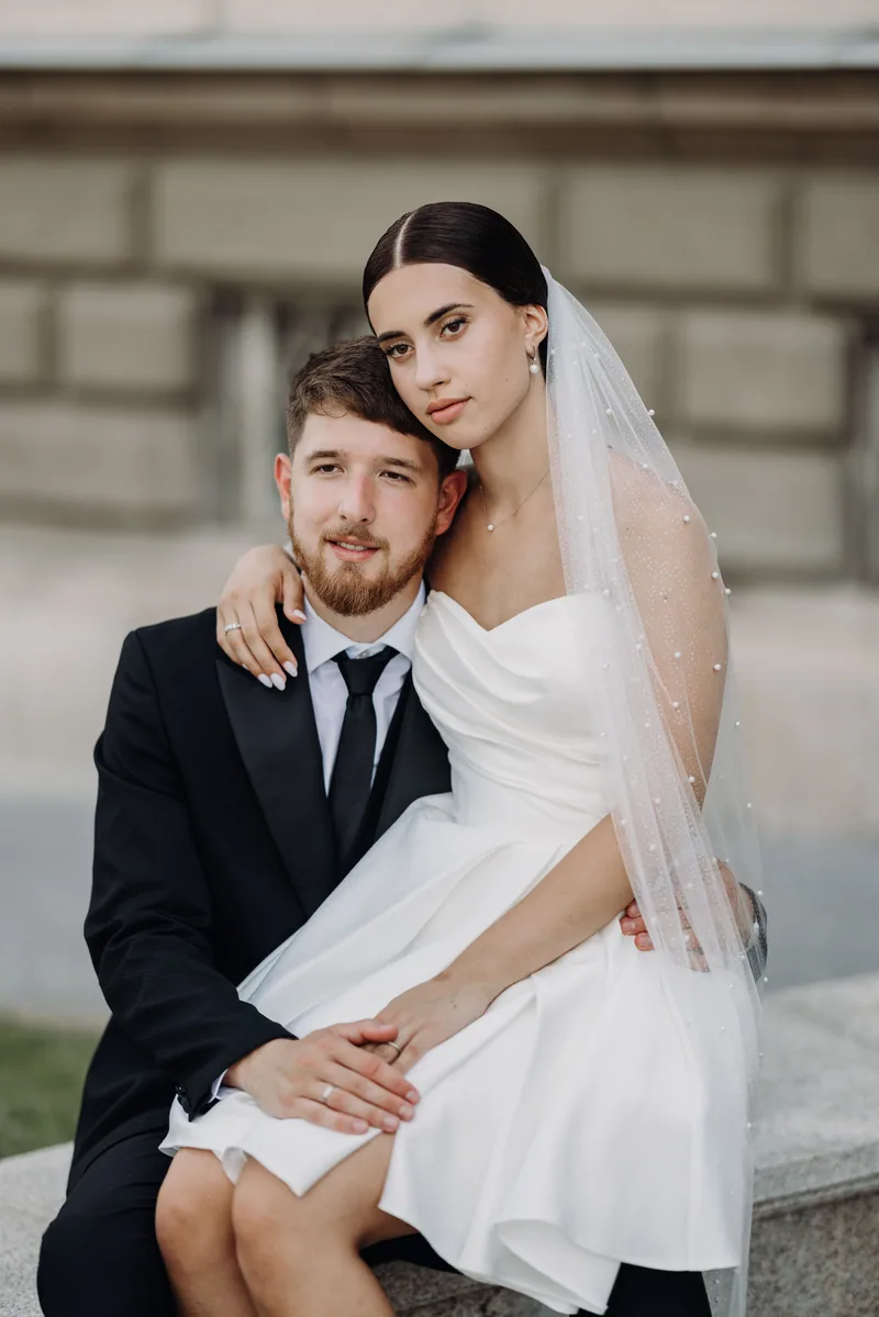 A bride sits on her groom's lap on a stone ledge, both looking at the camera with hands gently clasped.