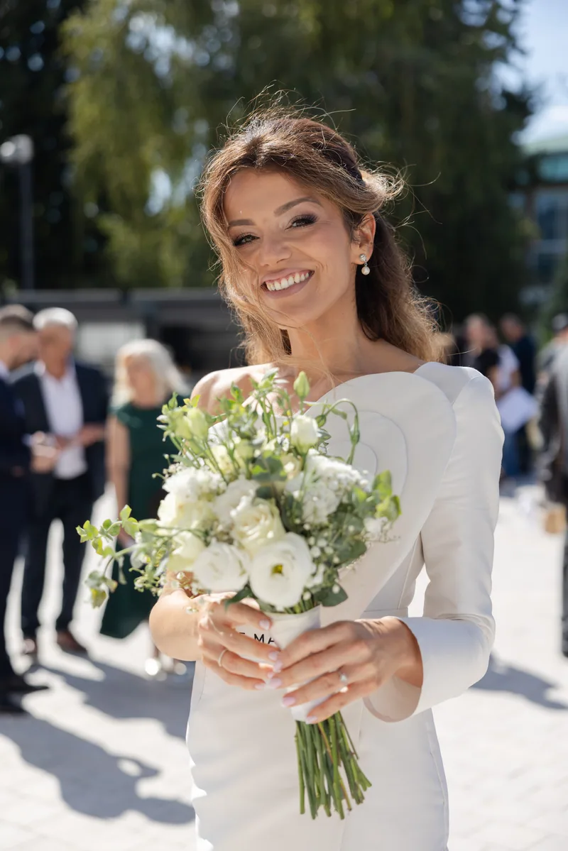 Sunlit bride smiles while holding a white and green bouquet outside the venue.