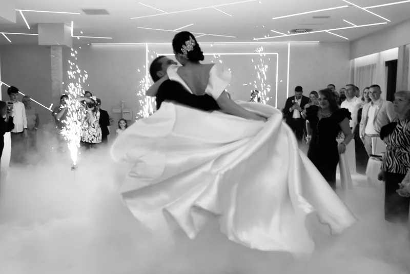 In black and white, a groom spins his bride so her gown flares wide through low fog before watching guests.
