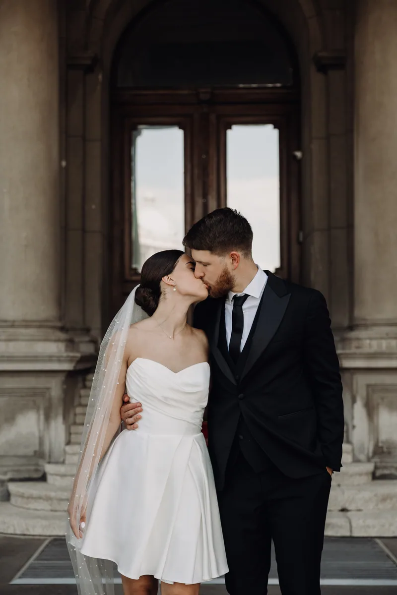 A bride and groom kiss standing in front of an ornate carved wooden door framed by stone columns.