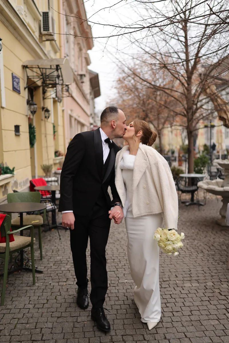 A couple kiss while walking down a quiet street lined with bare winter trees and cafe tables.