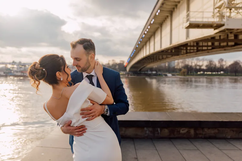 A couple embrace about to kiss on a riverside walkway at sunset with a lit bridge arcing over the river.