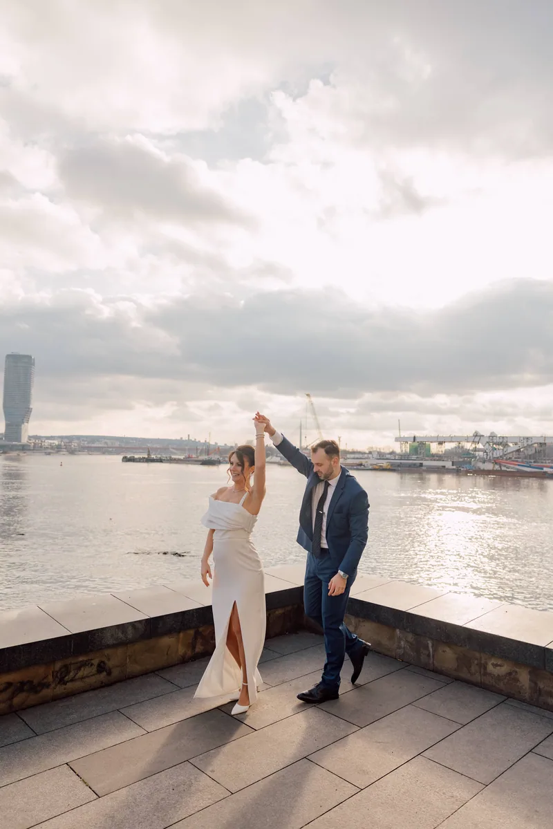 A groom twirls his bride on a riverside promenade at sunset beneath a wide cloudy sky over the water.
