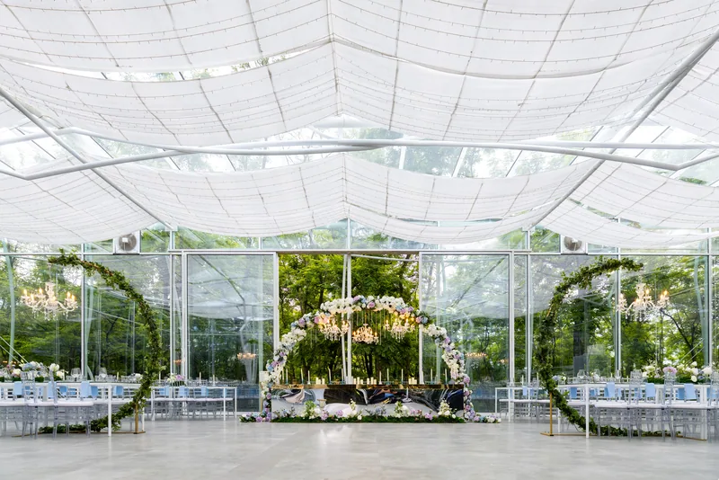 Wide symmetrical glass marquee reception with draped ceiling, floral arches and a central table