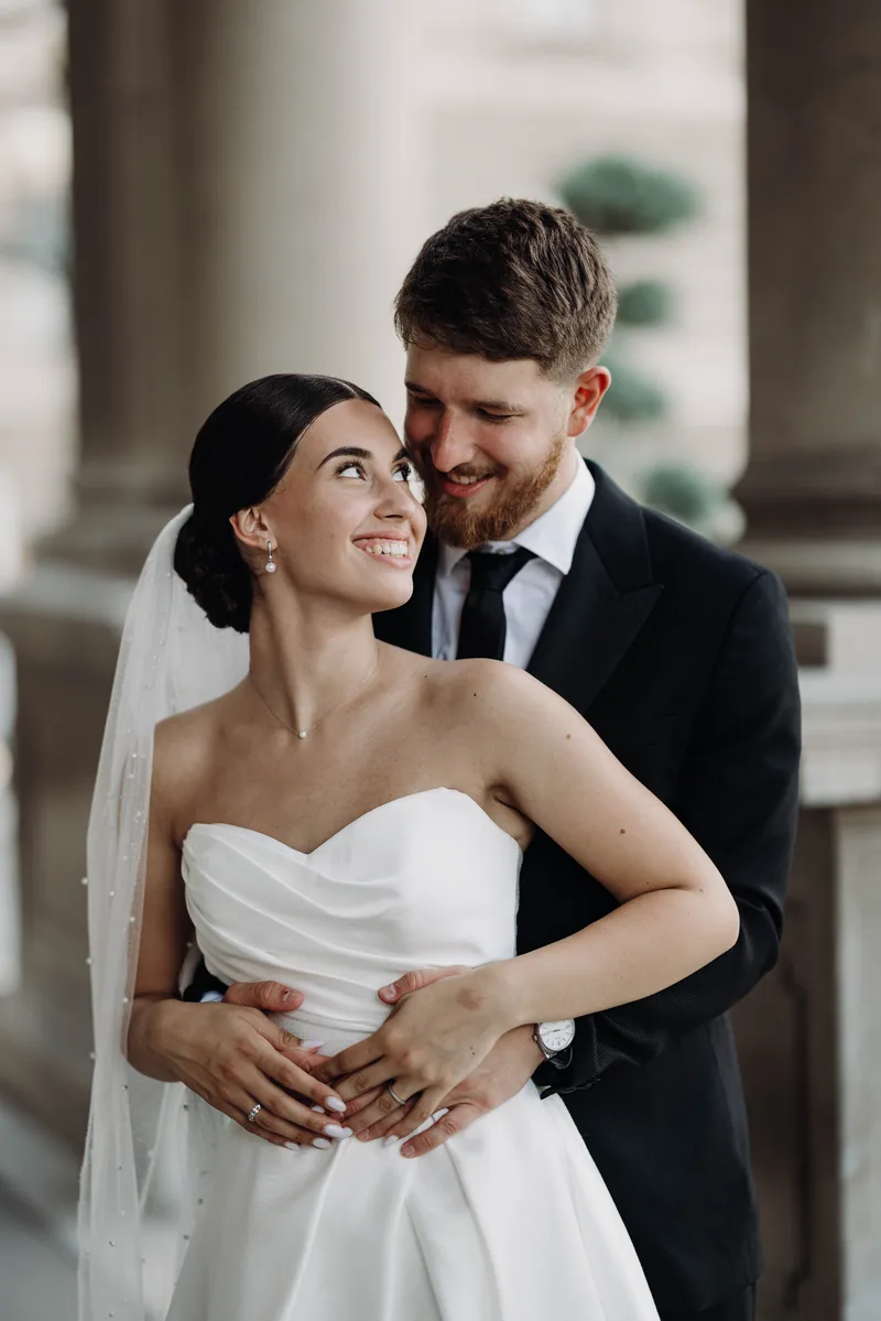 A groom embraces the bride from behind as they smile at each other, their hands clasped over her waist.
