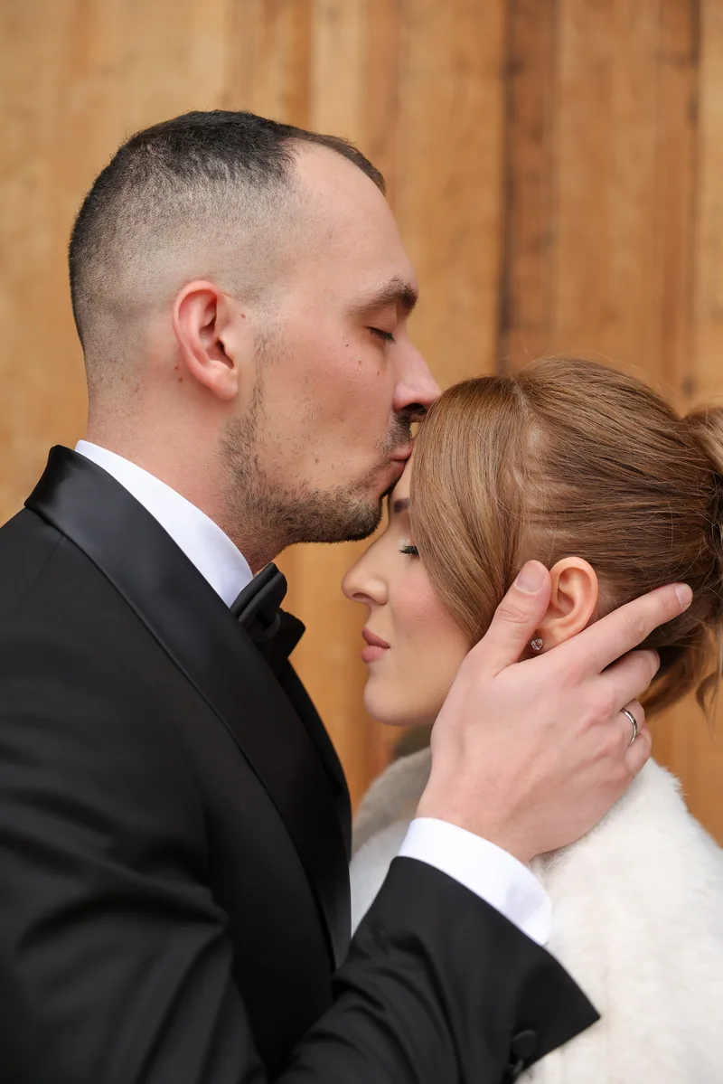 A groom in a tuxedo tenderly kisses his bride's forehead while cradling her face against a wooden wall.
