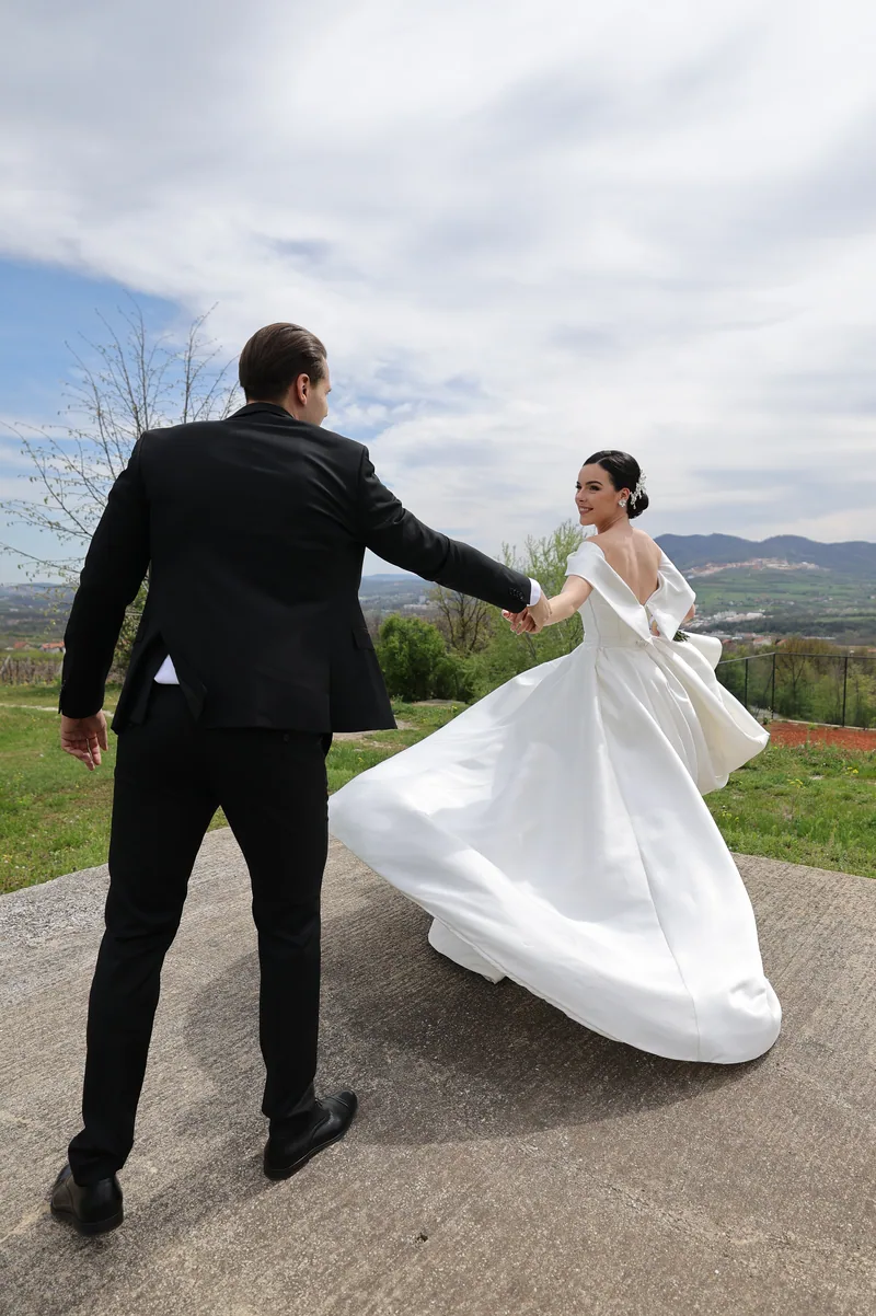 A groom leads his bride by the hand as her full ballgown swirls on a hilltop above a green valley.
