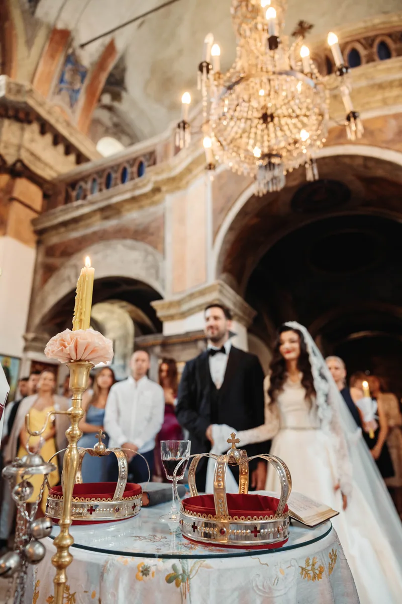 Wedding crowns and a candle on a table before an old frescoed church and crystal chandelier.