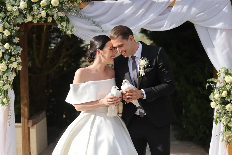 A bride and groom hold two white doves as they touch foreheads beneath a floral wedding arch.