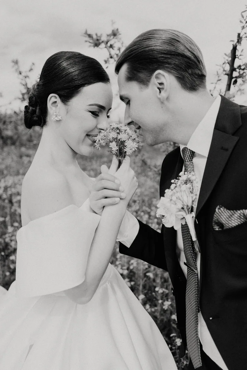 In black and white, a bride smells a small posy of wildflowers as her groom leans in close in a meadow.