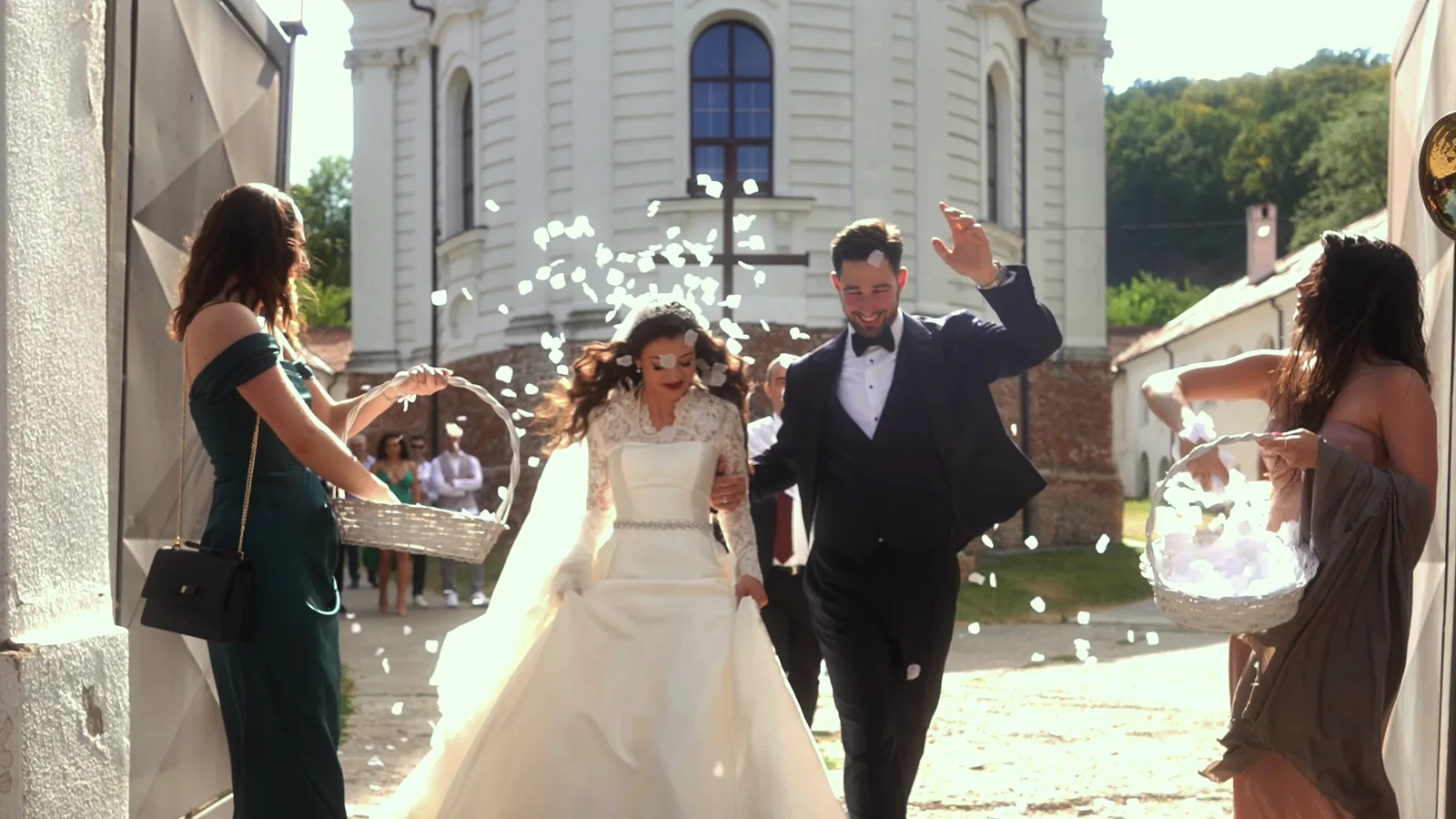 A bride and groom walk out of a church through a shower of petals thrown by guests.