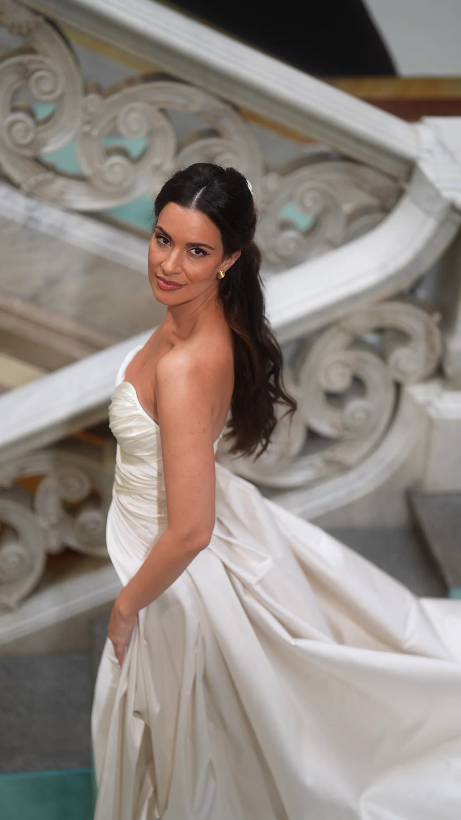 Une mariée en robe de satin se retourne par dessus son épaule sur un vaste escalier de marbre.